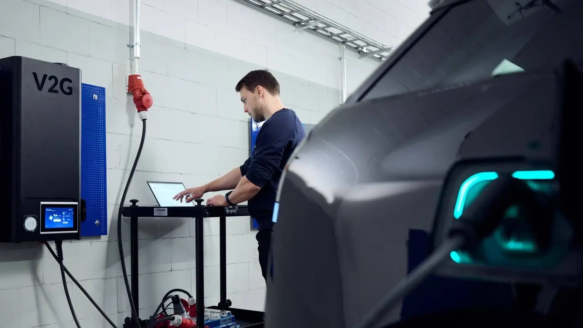 A man standing beside a laptop, V2G EV charger and an EV in a garage