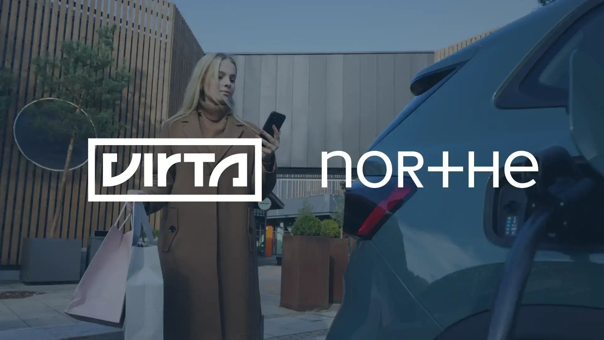 Kvinde holder en mobiltelefon ved en elbilslader med Virta- og Northe-logoobile phone next to an EV charging, with Virta and Northe logotypes
