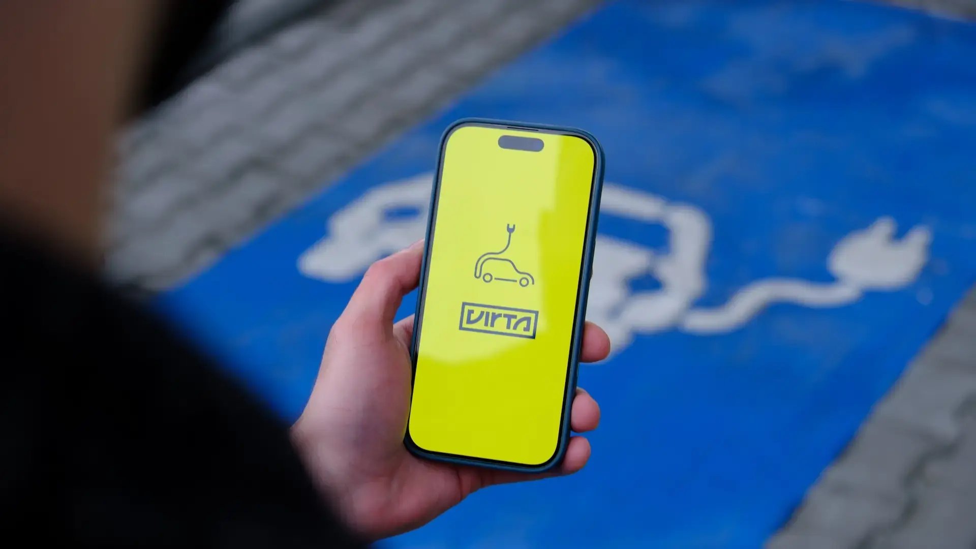 Man holding a smartphone with the Virta app in front of a car park with charging stations