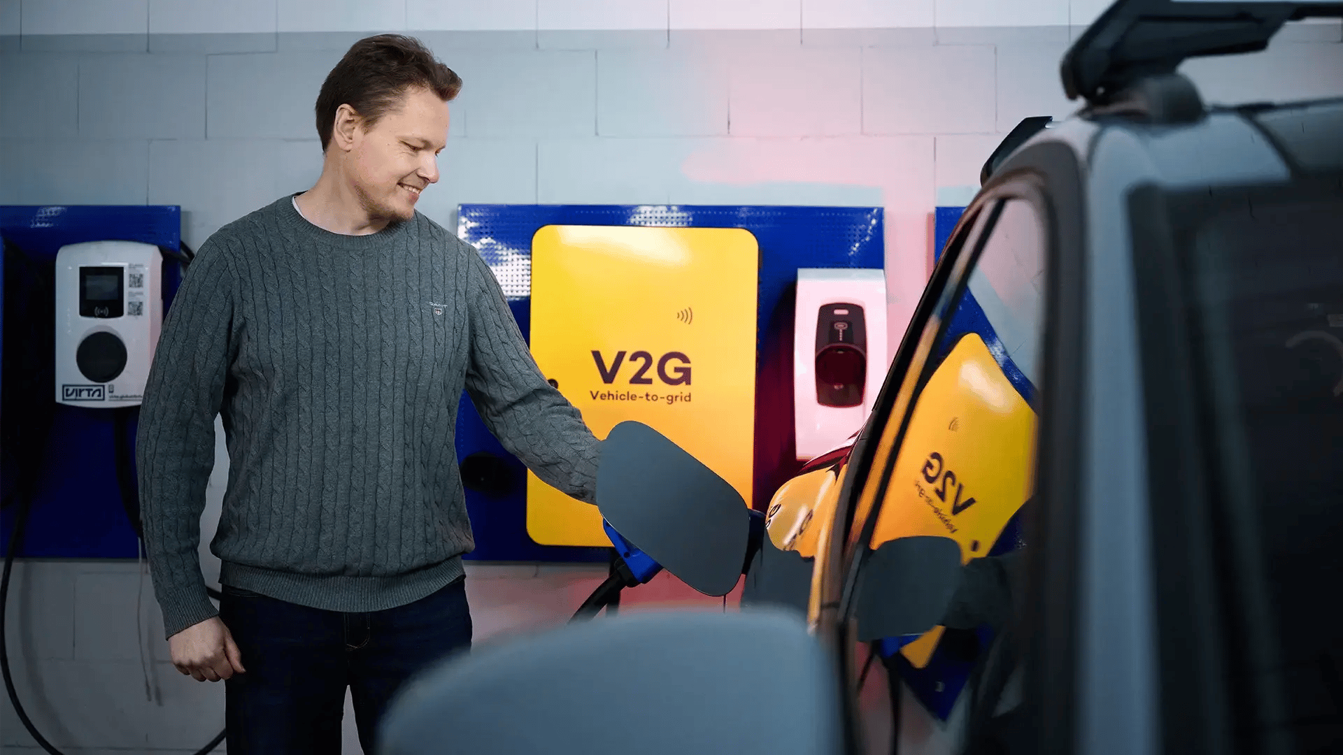 Man standing in a parking garage beside an EV, with an AC charger and V2G charger in the background