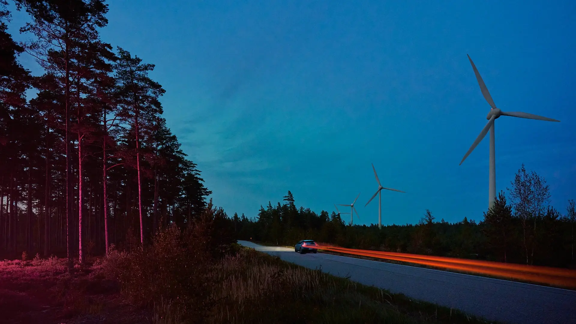 Auto elettrica che percorre una strada di campagna al crepuscolo attraverso una foresta con una turbina eolica sullo sfondo